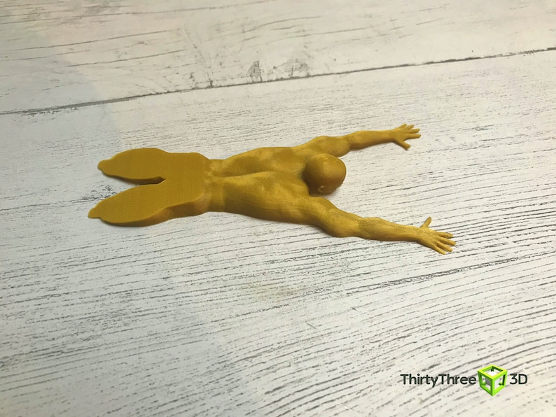 May include: A yellow 3D printed figurine of a person lying on their stomach with their arms outstretched. The figurine is on a white wooden surface.