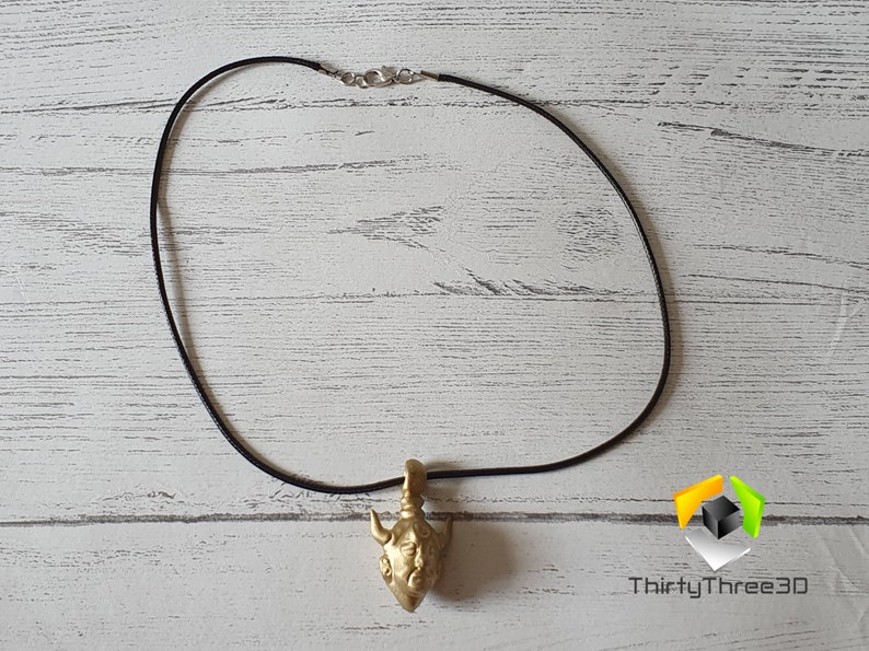 May include: A gold-colored pendant necklace with a detailed bull head design. The necklace is on a black cord.