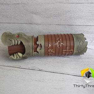 May include: A brown and gold 3D printed prop with a dragon head and intricate details. The prop has a red eye and is decorated with symbols and patterns.