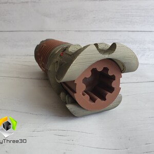 May include: A 3D printed model of a lightsaber hilt with a brown and grey colour scheme. The hilt is open, revealing a brown gear inside.