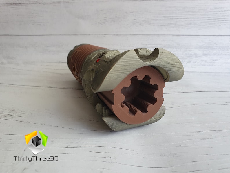 May include: A 3D printed model of a lightsaber hilt with a brown and grey colour scheme. The hilt is open, revealing a brown gear inside.