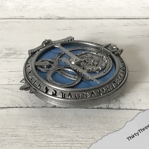 May include: A silver-colored metal pendant with a blue circular design and runes around the edge. The pendant features a gear-like mechanism with a silver-colored needle.