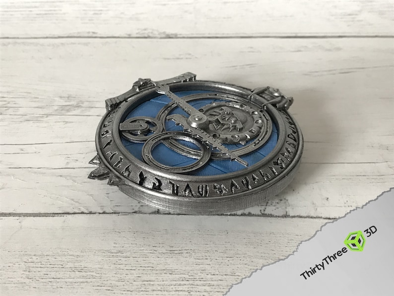 May include: A silver-colored metal pendant with a blue circular design and runes around the edge. The pendant features a gear-like mechanism with a silver-colored needle.