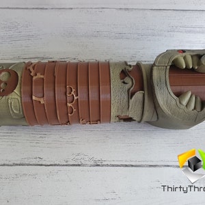 May include: A 3D printed, brown and green, detailed, articulated, dragon-like hand with a red eye. The hand is open and has sharp teeth.
