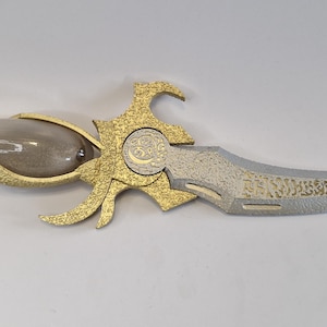 May include: An ornate dagger with a clear glass handle filled with a sand-like substance. The hilt and decorative elements are gold-colored, and the blade is silver with etched details. The end of the handle has a red accent.