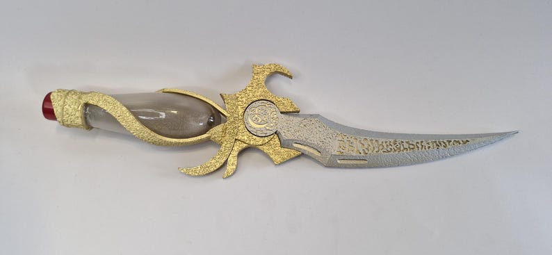 May include: An ornate dagger with a clear glass handle filled with a sand-like substance. The hilt and decorative elements are gold-colored, and the blade is silver with etched details. The end of the handle has a red accent.