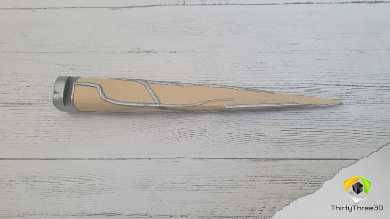 Op de afbeelding: Een beige en zilverkleurig dolk met een getextureerd oppervlak. De dolk heeft een puntige punt en een afgeronde handgreep.