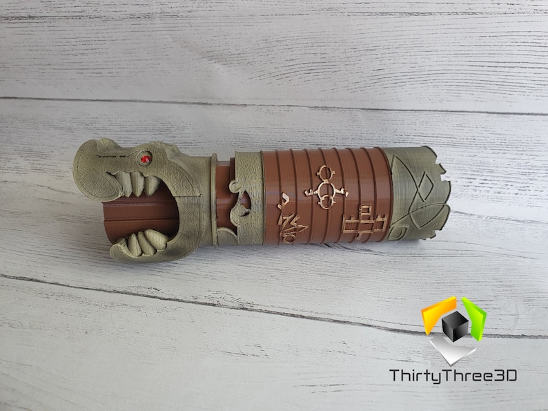 May include: A 3D printed brown and gold-colored prop resembling a mythical creature with a red eye and sharp teeth. The prop has intricate details and carvings, including a series of symbols and a dragon-like head.