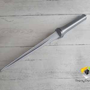 May include: A silver-colored dagger with a pointed tip and a handle. The dagger is lying on a white wooden surface.