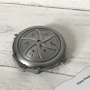 May include: A silver-coloured metal disc with a circular design and symbols. The disc has a raised centre and a textured surface.