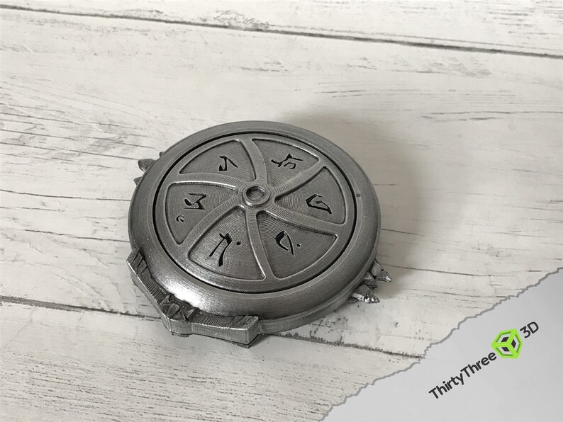 May include: A silver-coloured metal disc with a circular design and symbols. The disc has a raised centre and a textured surface.