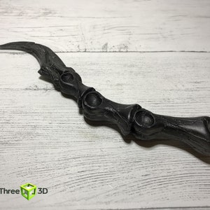 May include: A black, curved wand with a bone-like design. The wand has a textured surface and features a series of rounded, open sections along its length. The wand is lying on a white wooden surface.