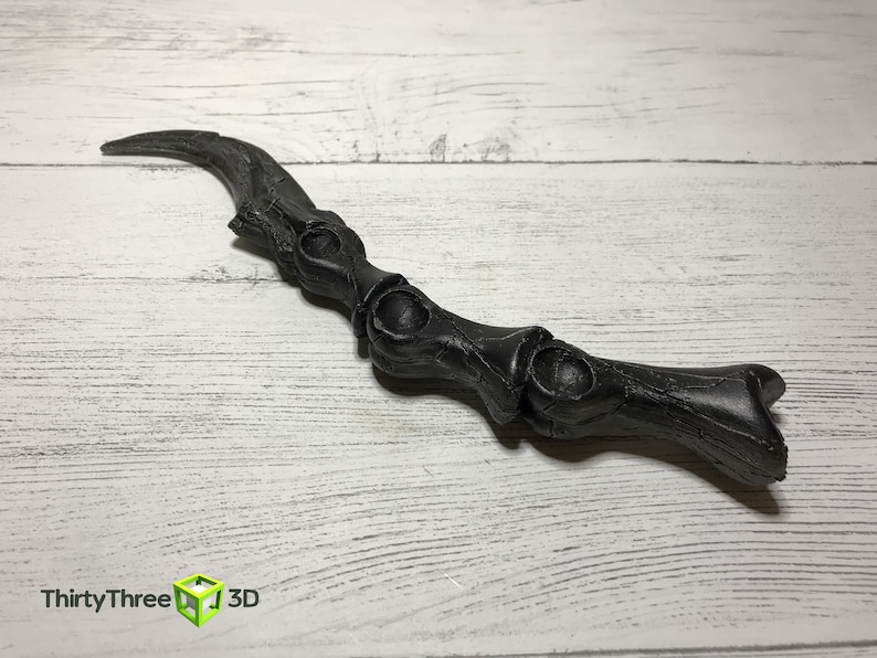 May include: A black, curved wand with a bone-like design. The wand has a textured surface and features a series of rounded, open sections along its length. The wand is lying on a white wooden surface.