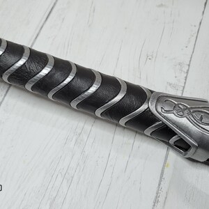 May include: Close-up of a black and silver handle with a leather grip and a silver metal cap with a decorative design. The handle is for a prop weapon.