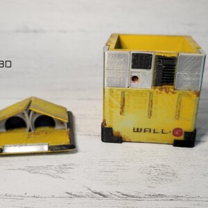 May include: A yellow, cube-shaped box with a rusty, weathered look. The box has a removable lid and the text "WALL-E" printed on the front. The lid is open and shows the inside of the box.