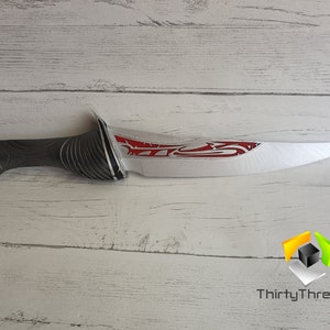 May include: A silver and red fantasy-style dagger with a black handle. The blade has a red design on it. The dagger is on a white wooden surface.