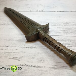 May include: A brown, detailed, decorative sword with a textured handle and a blade with intricate carvings. The sword is lying on a white wooden surface.