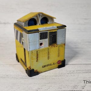 May include: A yellow and gray WALL-E robot toy with a red circle on the front. The robot has a small, rectangular body with a square head and two large, round eyes.