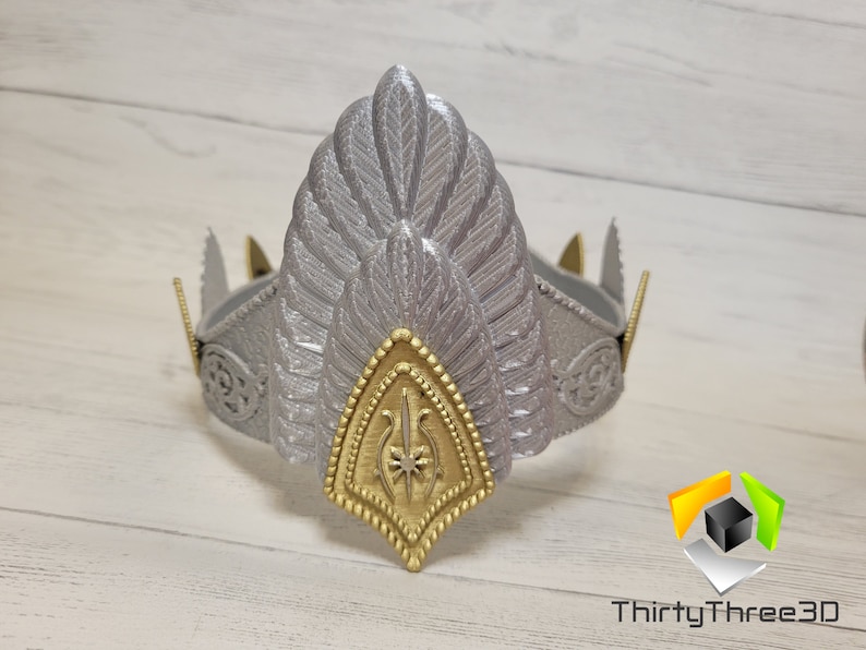 May include: A silver and gold crown with a feathery design. The crown has a gold center piece with a star and a stylized design.