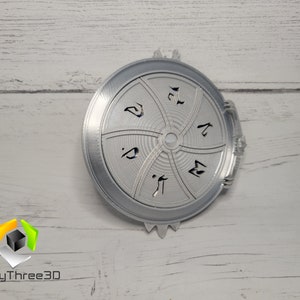 Puede incluir: Un disco de metal plateado con un dise&ntilde;o circular y cinco secciones. Cada secci&oacute;n tiene un s&iacute;mbolo o dise&ntilde;o diferente. El disco tiene un borde elevado y una peque&ntilde;a manija en el lateral.