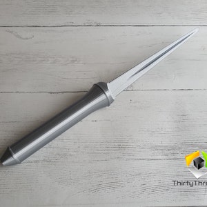 May include: A silver-colored, long, thin, pointed object with a handle. The object is lying on a white wooden surface.