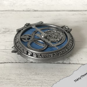 May include: A silver-coloured metal pendant with a blue circular design and runes around the edge. The pendant features a gear-like mechanism with a silver-coloured needle.