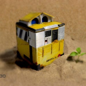 May include: A yellow and black robot toy with a white panel and the word "WALL-E" printed on the side. The robot is sitting on a sandy surface.