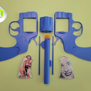 Op de afbeelding: Een gedemonteerde blauwe speelgoedrevolver met een oranje punt, met twee framehelften, een cilinder en andere componenten. De afbeelding bevat ook twee vintage pin-up afbeeldingen.