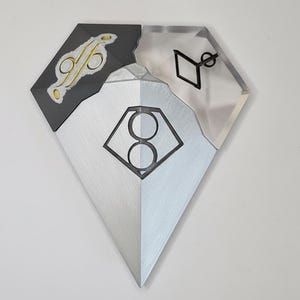 Pu&ograve; includere: Un'opera d'arte da parete a forma di diamante con un design geometrico. L'opera &egrave; divisa in tre sezioni: una superficie metallica argentata, una sezione nera con accenti dorati e una sezione trasparente con una forma geometrica nera.