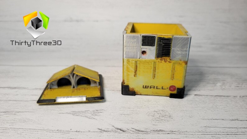 May include: A yellow, cube-shaped box with a rusty, weathered look. The box has a black and gray panel on the front with the word "WALL-E" printed on it. The box is open and the lid is separate. The lid is also yellow and has a design that resembles a robot's head.