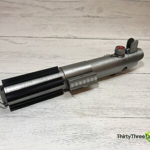 May include: A silver lightsaber prop with a black handle and a red button on the top. The lightsaber has a row of small white lights on the side.