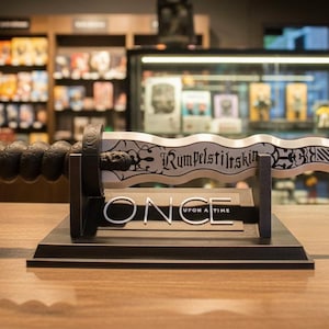 May include: A decorative dagger with a black handle and a silver blade, displayed on a black stand. The blade is inscribed with the words "Rumpelstiltskin" and the stand reads "ONCE UPON A TIME". The dagger is approximately 15 inches long.