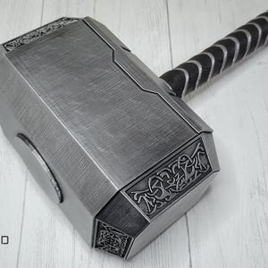 May include: A silver-colored Mjolnir hammer with a black leather handle. The hammer head has intricate details and a textured surface.
