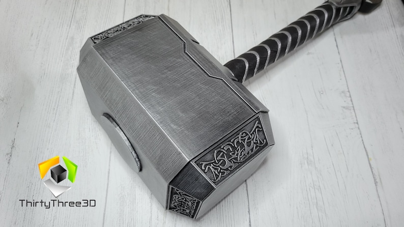 May include: A silver-colored Mjolnir hammer with a black leather handle. The hammer head has intricate details and a textured surface.