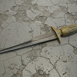 May include: A decorative dagger with a detailed, patterned blade and a gold-colored handle. The handle features an ornate design and a red gem. The dagger is lying on a cracked, gray surface.