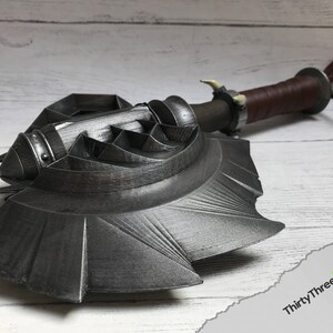 May include: A 3D printed, black metal axe with a detailed, textured design. The axe has a long handle with a brown leather wrap and a metal cap. The axe head is shaped like a crescent moon with a sharp edge.