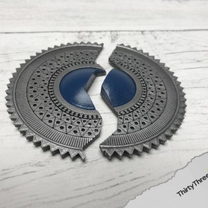 May include: A silver-colored, circular, decorative piece with a blue center. The piece is split into two halves, each with a detailed, geometric pattern.