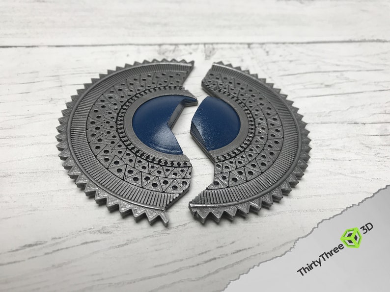 May include: A silver-colored, circular, decorative piece with a blue center. The piece is split into two halves, each with a detailed, geometric pattern.