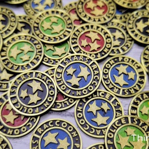 Puede incluir: Primer plano de un mont&oacute;n de fichas de metal de color dorado con un dise&ntilde;o en blanco y negro. Las fichas muestran las palabras "Police Stars Raccoon" y tres estrellas. Las fichas est&aacute;n dispuestas en un patr&oacute;n aleatorio.