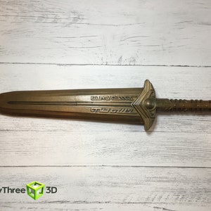 May include: A gold-colored sword with intricate details and a textured handle. The sword is lying on a white wooden surface.