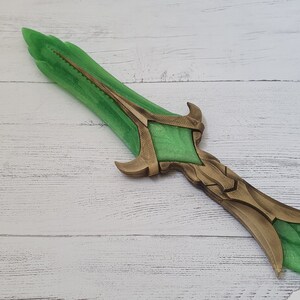 May include: A green and gold fantasy dagger with a detailed design. The blade is green and the hilt is gold with intricate details.