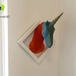 May include: A 3D printed unicorn head sculpture in a white frame. The unicorn is made of orange, blue, and green plastic. The sculpture is mounted on a white wall.