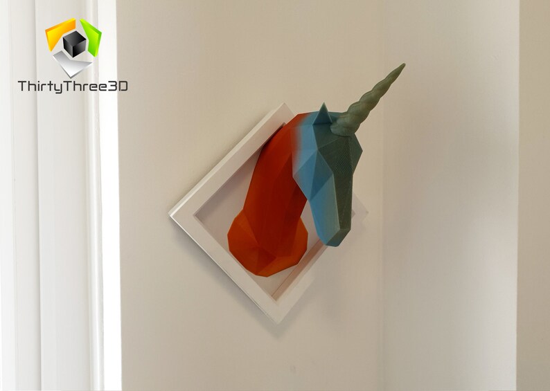May include: A 3D printed unicorn head sculpture in a white frame. The unicorn is made of orange, blue, and green plastic. The sculpture is mounted on a white wall.