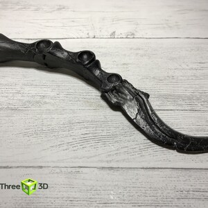 May include: A black, claw-shaped object with a textured surface, possibly 3D-printed. The design features a series of rounded protrusions and a curved blade-like end. The object is set against a light wood-grain background.