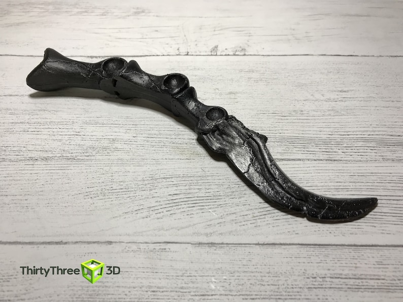 May include: A black, claw-shaped object with a textured surface, possibly 3D-printed. The design features a series of rounded protrusions and a curved blade-like end. The object is set against a light wood-grain background.