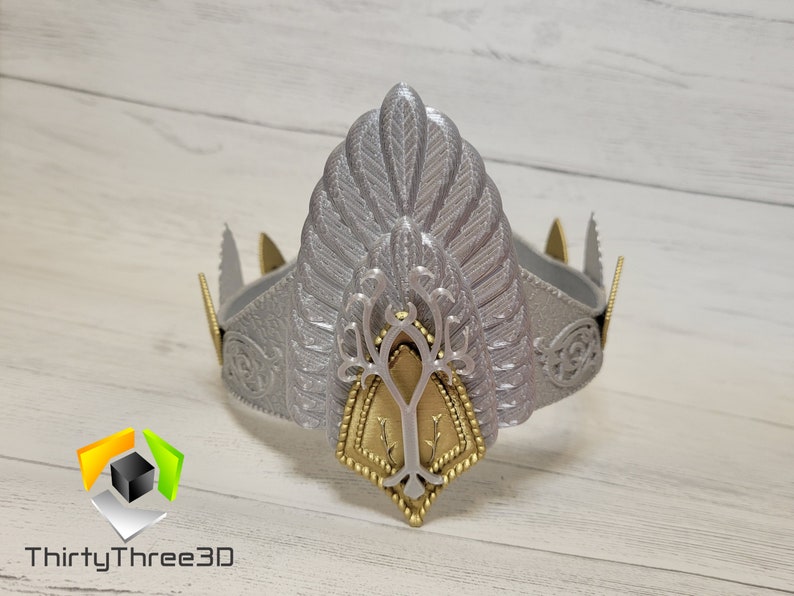 May include: A silver crown with a gold tree design in the center. The crown has a feathery design on the top and a textured surface.