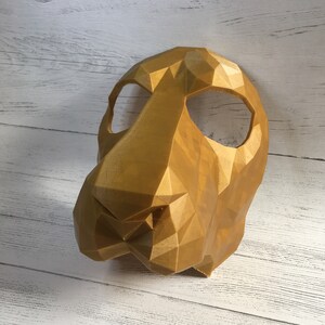 May include: A gold-colored, 3D printed mask with a geometric design. The mask has two eye holes and a prominent nose.