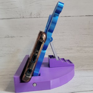 Peut inclure: Un support de t&eacute;l&eacute;phone violet avec un support de t&eacute;l&eacute;phone bleu. Le support a une petite plateforme avec des marches et une base circulaire plus grande. Le t&eacute;l&eacute;phone est dans un &eacute;tui marron.
