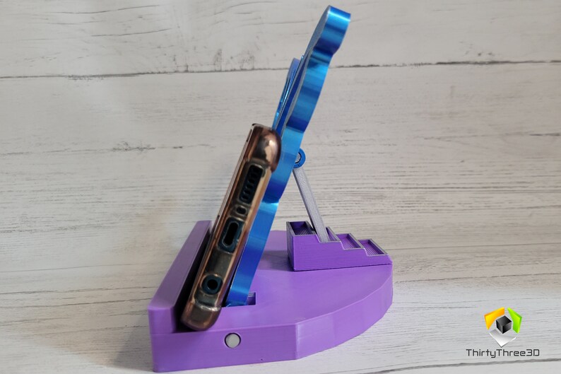 Peut inclure: Un support de t&eacute;l&eacute;phone violet avec un support de t&eacute;l&eacute;phone bleu. Le support a une petite plateforme avec des marches et une base circulaire plus grande. Le t&eacute;l&eacute;phone est dans un &eacute;tui marron.