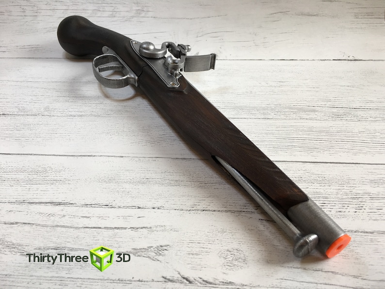 Puede incluir: Una r&eacute;plica de pistola de chispa con culata de madera oscura y componentes met&aacute;licos. La pistola tiene gatillo, martillo y ca&ntilde;&oacute;n. El extremo del ca&ntilde;&oacute;n tiene una punta naranja. La imagen tambi&eacute;n incluye el texto "ThirtyThree 3D".